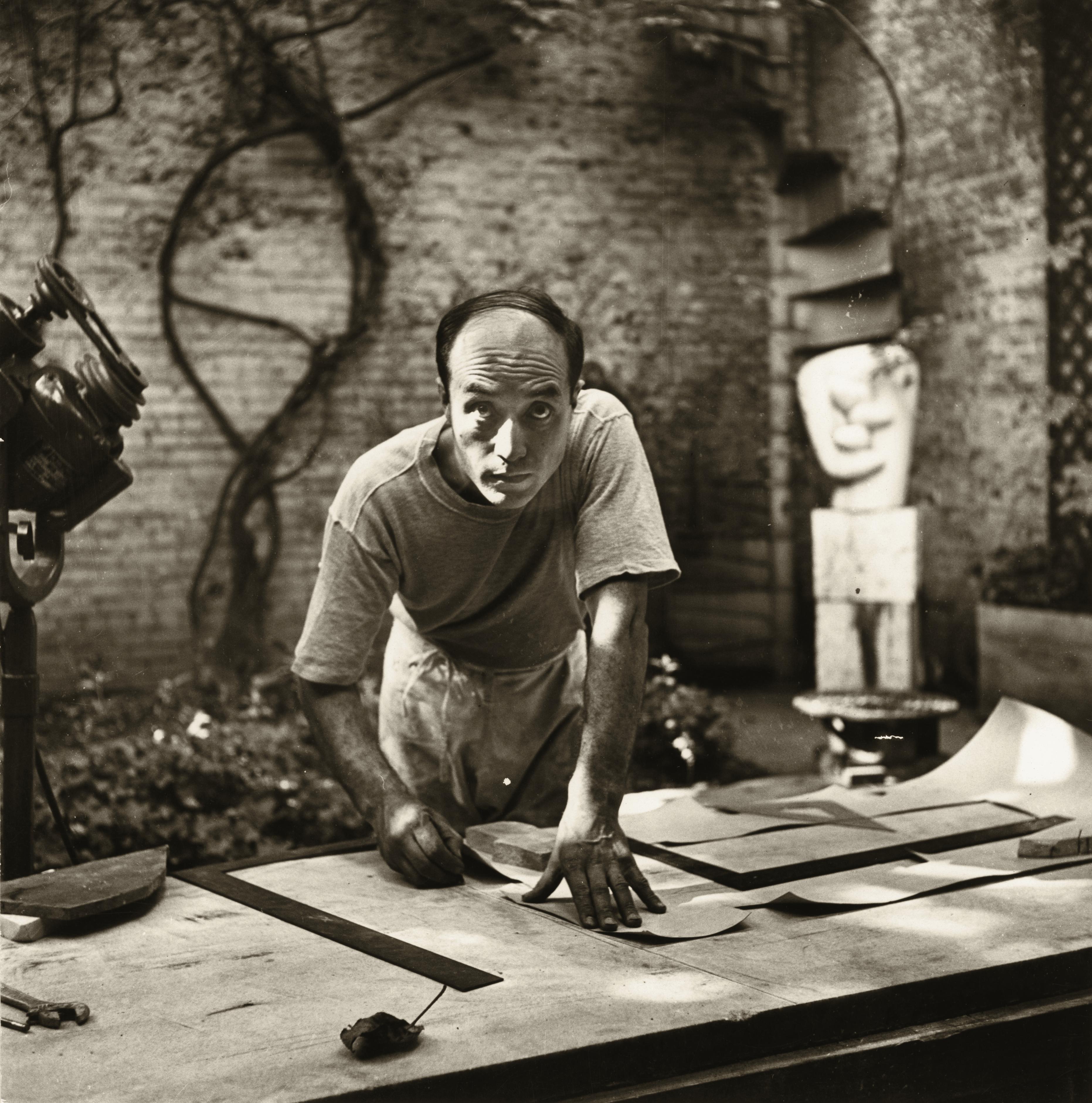 Black-and-white photograph of a man leaning over a worktable in an outdoor studio, carefully shaping or tracing paper or stone. Sculptural tools and materials surround him, with abstract sculptures and a brick wall visible in the background, conveying a focused moment of artistic work.