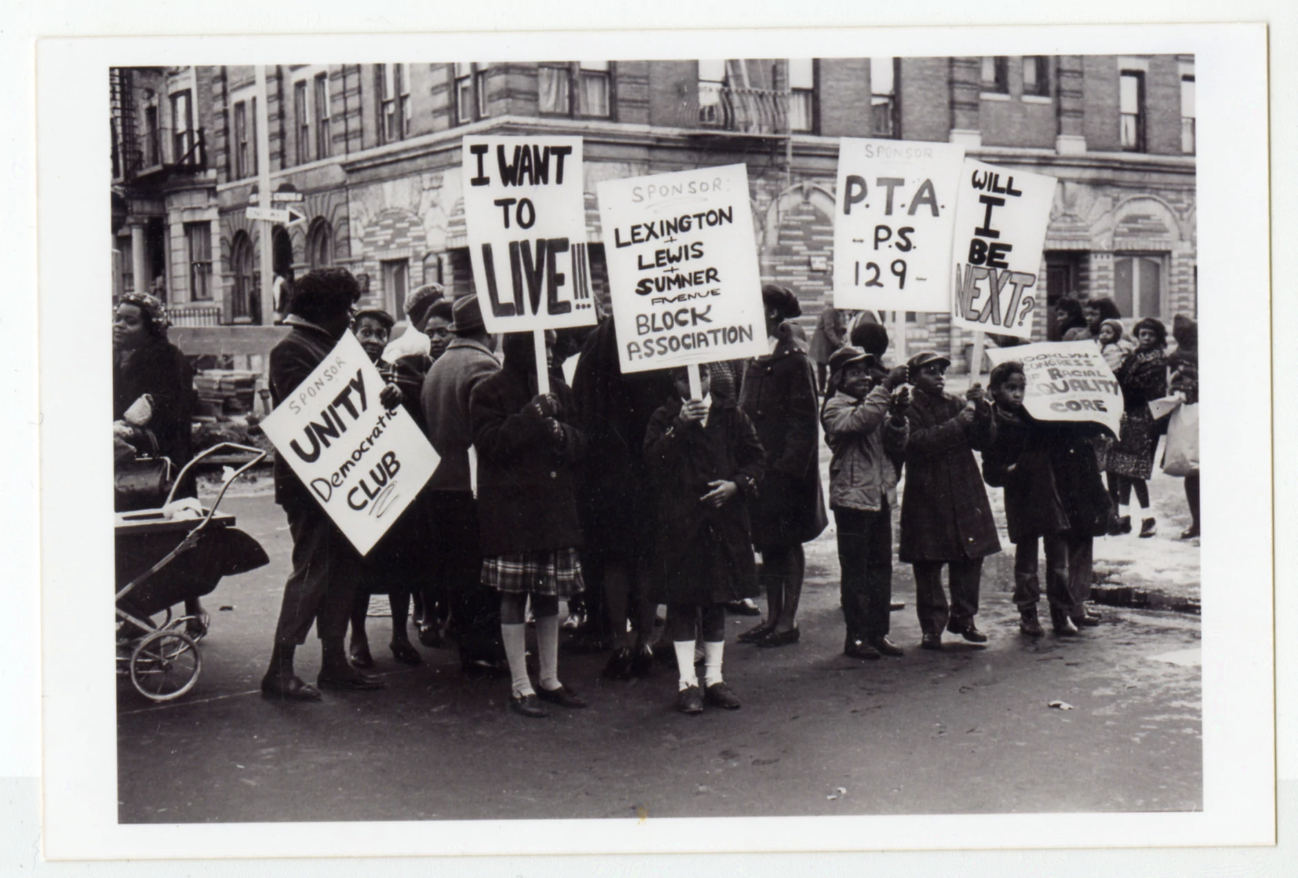 A group of adults and children protest police brutality. Children hold picket signs that read: "Sponsor Unity Democratic Club" "I want to live!!!" "Sponsor Lexington + Lewis + Sumner Avenue Block Association" "Sponsor PTA PS 129" "Will I be next?" "Brooklyn Congress of Racial Equality CORE"