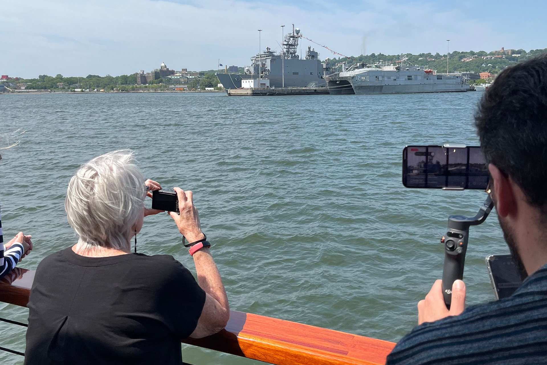 Two people with cameras on a boat taking photos of a military navy ship in NY Harbor