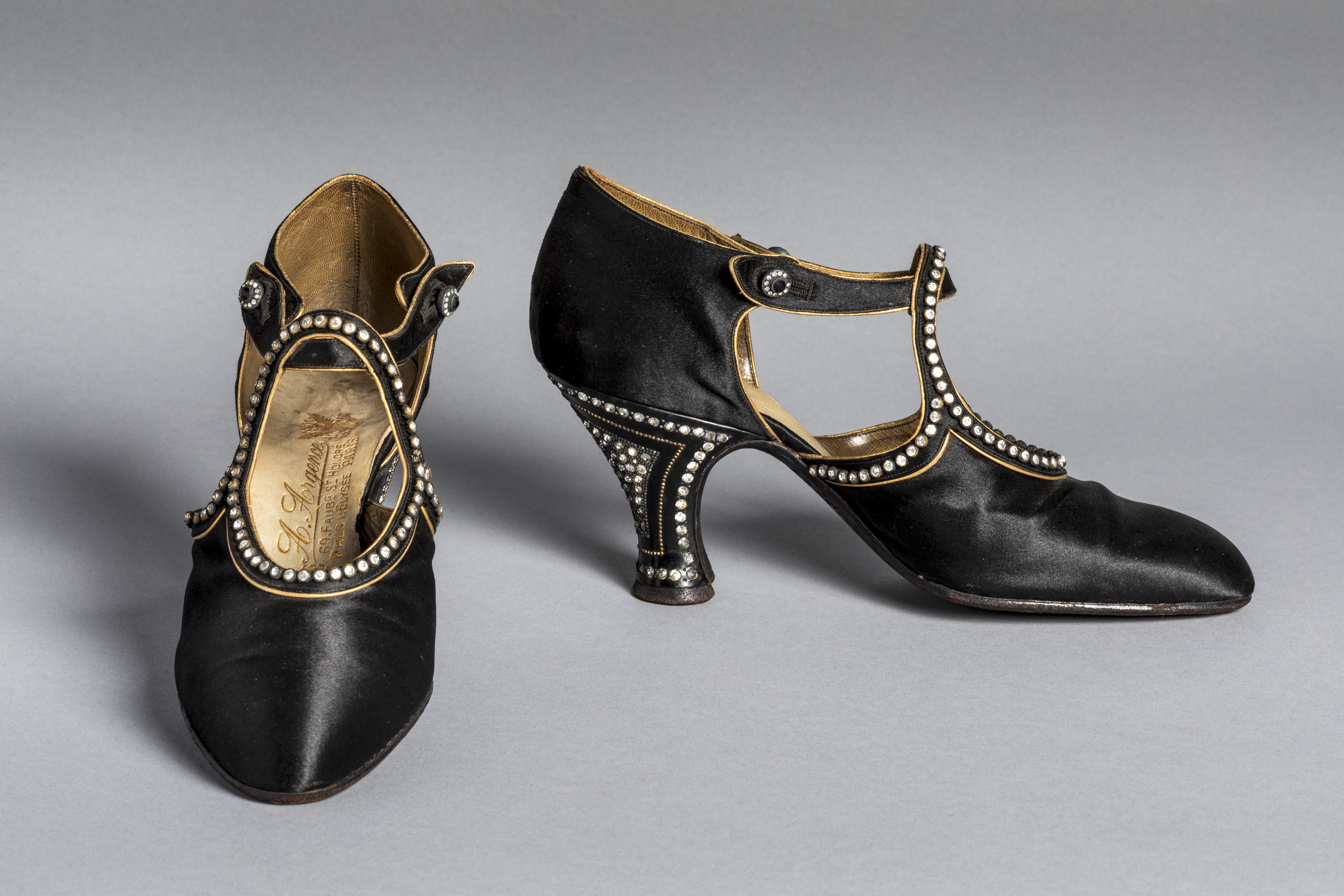A pair of elegant black satin women's heels from the early 20th century, featuring T-straps and detailed with rows of small pearls along the edges and around the flared heels. The shoes have an Art deco design with button closures and gold lining visible inside.