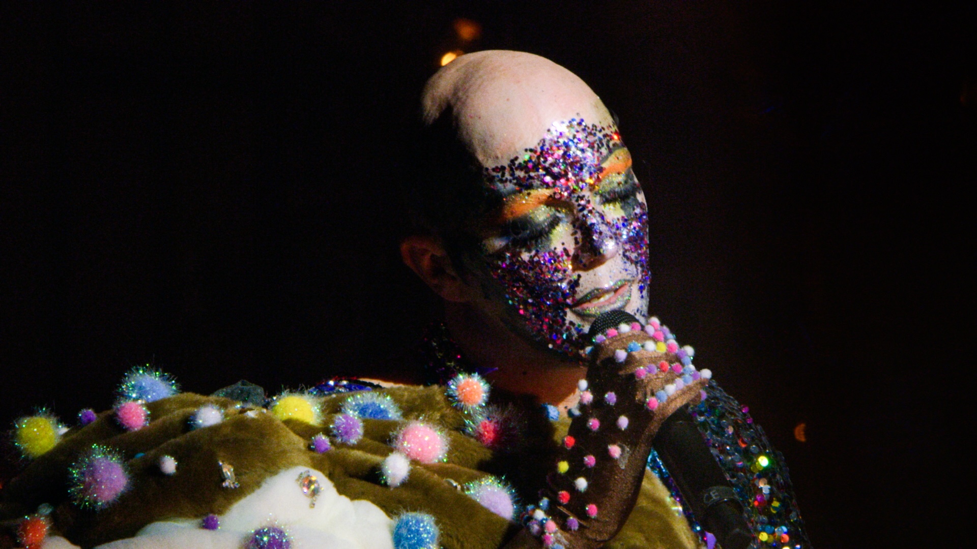Taylor Mac is shown from shoulders up holding a microphone. Taylor's face is covered in glitter, with orange eyebrows and green eyeshadow. Sparkly pom poms are affixed to Taylor's gloved hands and furry taupe costume.