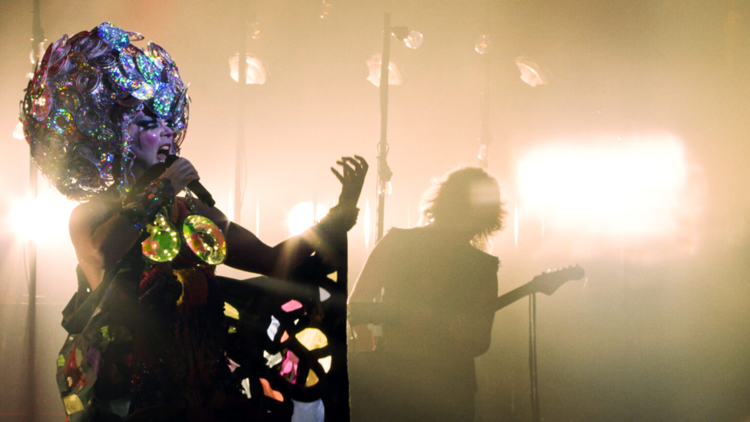Taylor Mac is pictured on the left of the shot holding in elaborate drag. Taylor is singing into a microphone with one hand outstretched, mouth wide and eyes intense. Taylor wears a large sparkly headpiece, evoking a discoball, made of sparkling disks. The silhouette of a guitar player can be seen in the background.