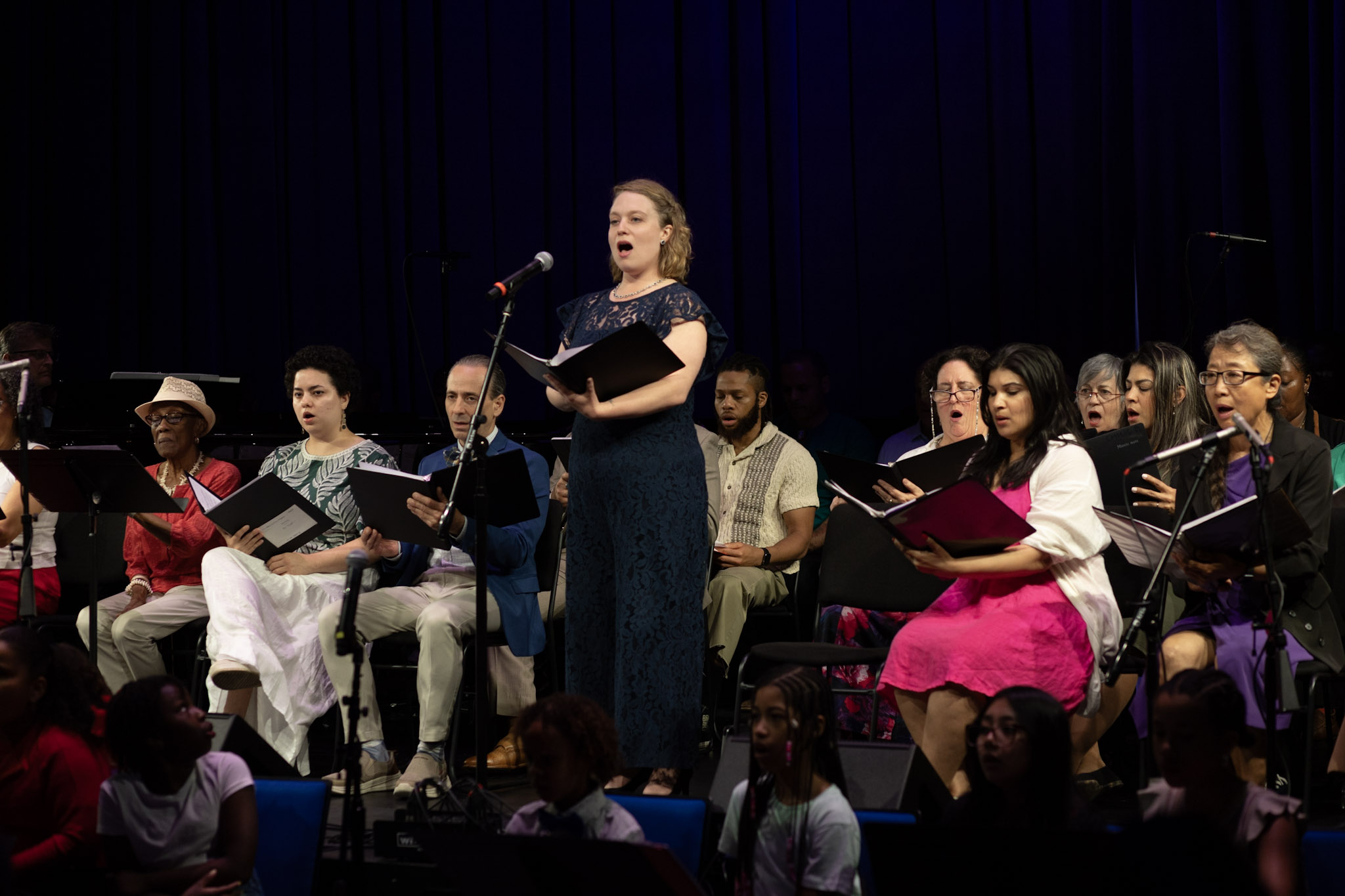 Soloist in navy dress standing at mic accompanied by a seated, intergenerational, diverse, colorfully clad chorus