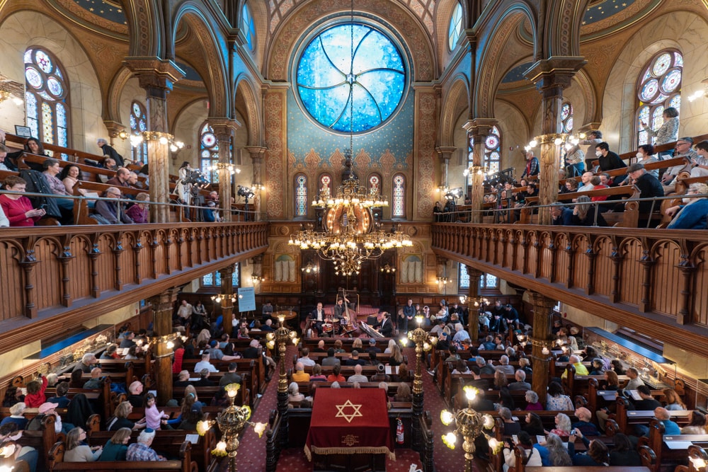 Historic synagogue with blue stained glass window filled with guests attending concert.