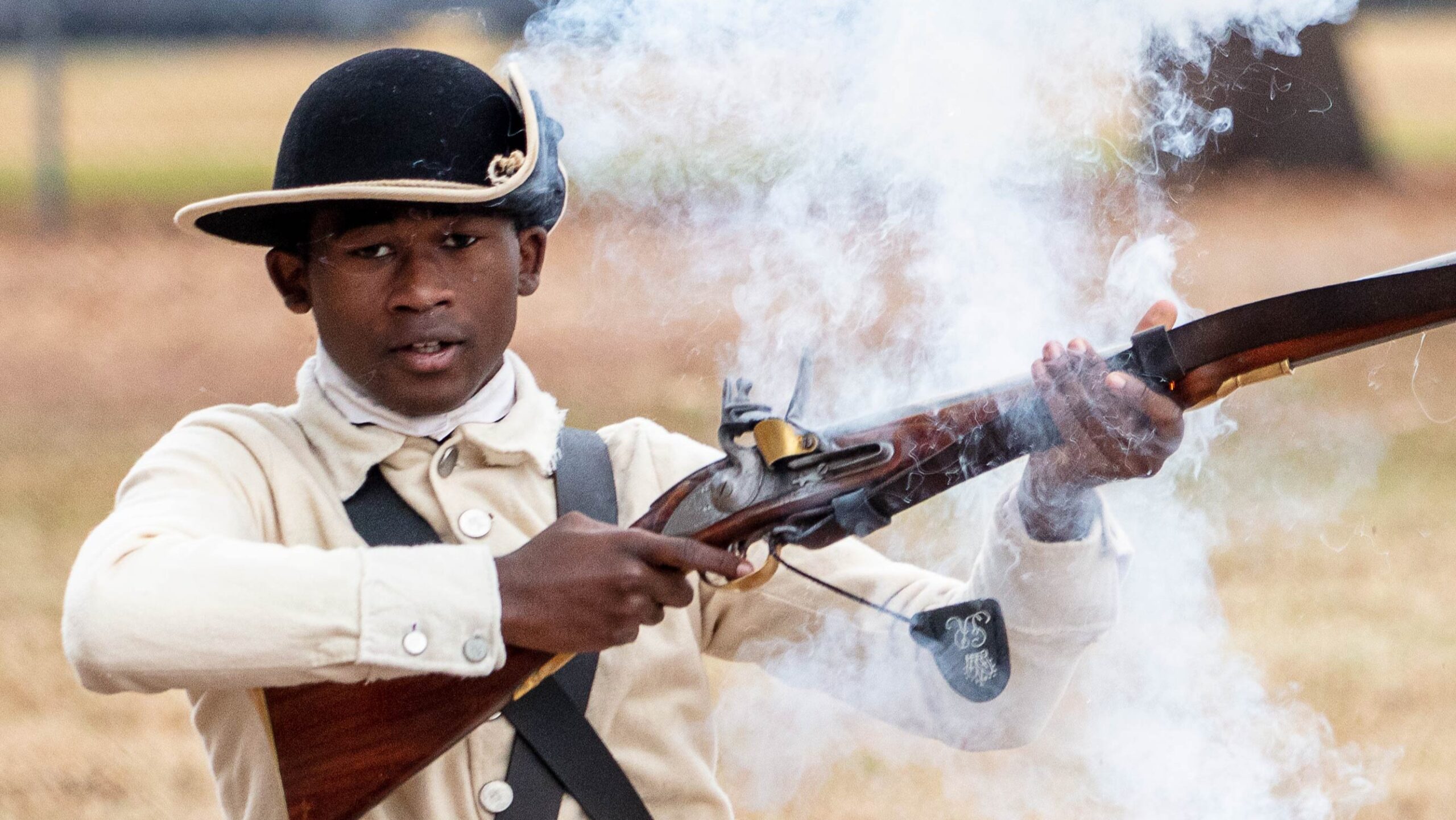 a young Black man dressed in Revolutionary War patriot clothes holds a musket that has just been fired and is smoking