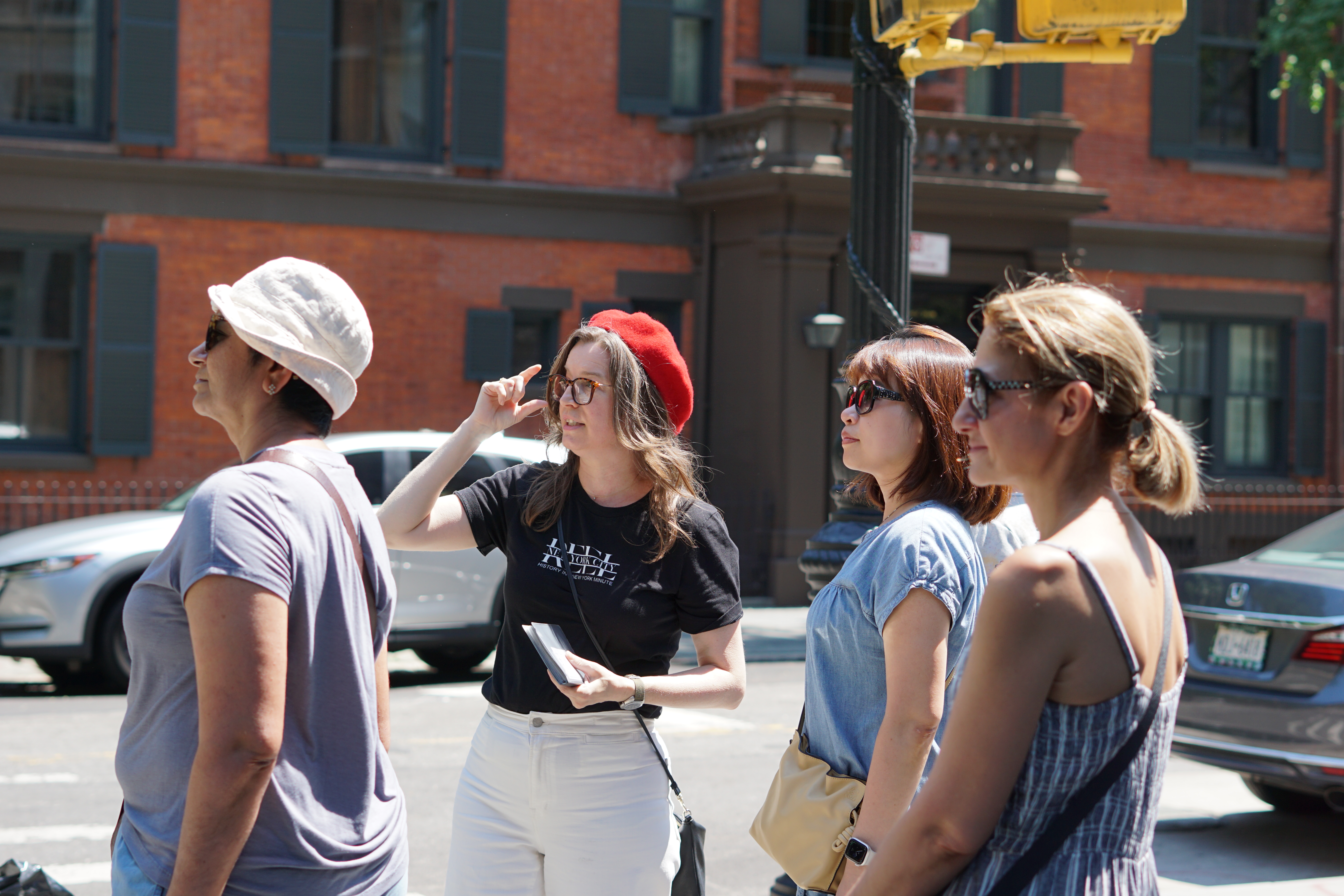 A group of four women standing on a city street, one wearing a red beret and pointing, with red brick building in the background.